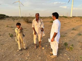 Three people—an elderly man, a young boy, and another adult man—stand in conversation on a dry, open landscape scattered with low shrubs. They wear traditional light-colored clothing and hold wooden walking sticks. In the background, multiple large wind turbines rise across the horizon under a partly cloudy sky, suggesting a juxtaposition of local community life and renewable energy development. A few goats can be seen grazing in the distance.