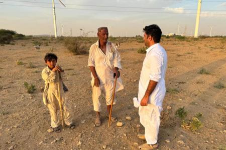 Three people—an elderly man, a young boy, and another adult man—stand in conversation on a dry, open landscape scattered with low shrubs. They wear traditional light-colored clothing and hold wooden walking sticks. In the background, multiple large wind turbines rise across the horizon under a partly cloudy sky, suggesting a juxtaposition of local community life and renewable energy development. A few goats can be seen grazing in the distance.